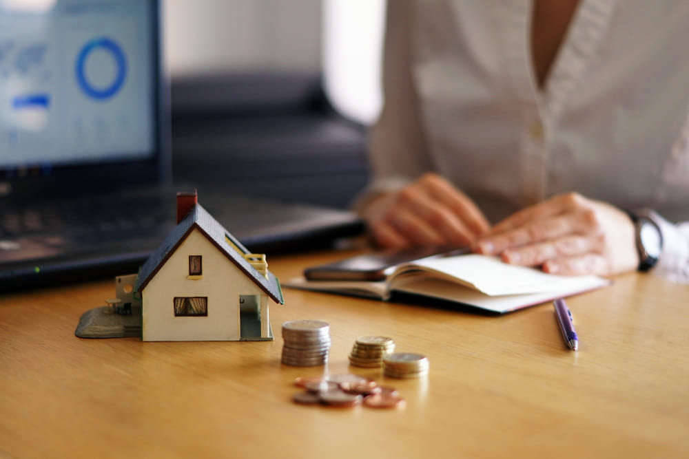 Posso vender um imóvel durante o inventário? Imagem em que consta uma miniatura de uma casa e moedas em cima de uma mesa, com uma pessoa sentada na cadeira com as mãos em cima de um bloco de notas.